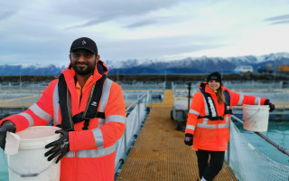 Two people hold large white buckets on a salmon farm