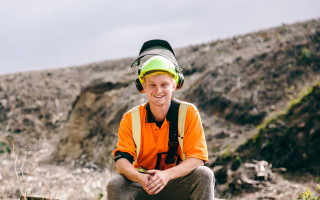 A man in a hard hat and a high vis vest sits on a log with chopped down trees around him